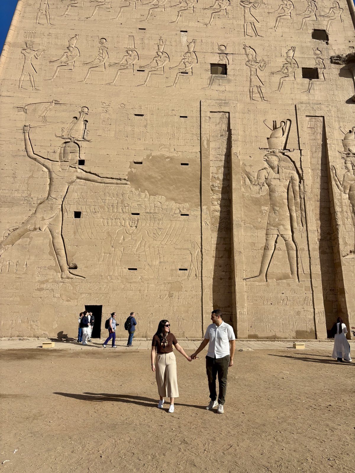 Moni y Adri ante la gran fachada del Templo de Edfu con relieves de dioses egipcios tallados en la piedra