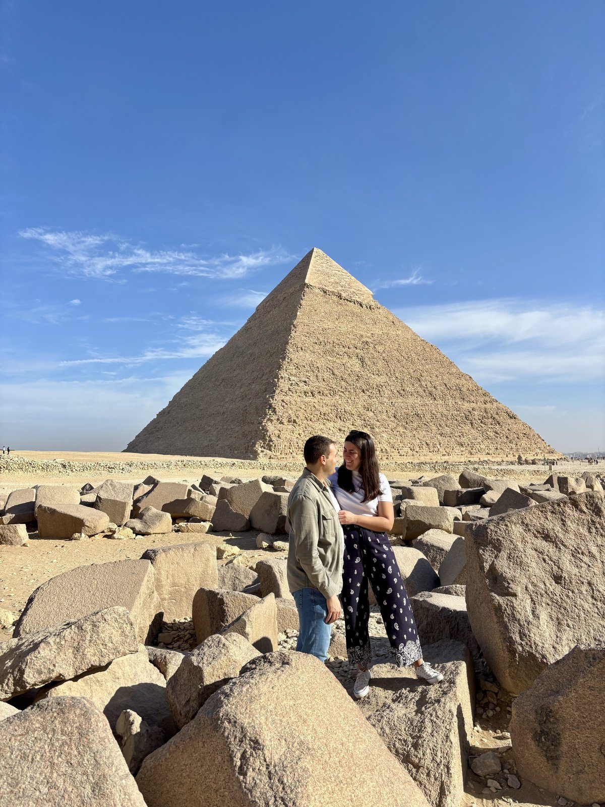 Moni y Adri mirándose entre bloques de piedra con la Pirámide de Kefrén de Guiza al fondo bajo cielo azul